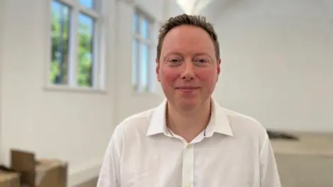 Tom Jackson/BBC Head and shoulder image of Matthew Webb. He has short red hair and is wearing a white shirt. He is standing in the Mill Road library building.
