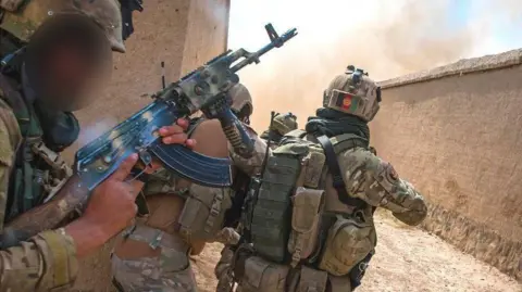 Ben Taggart Three Afghan soldiers in camouflage gear with guns peer around the corner of a sand-colored building, with smoke seen around the corner.