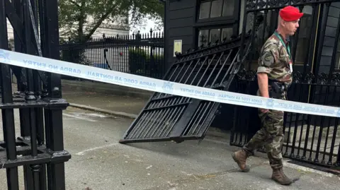 PA Media Military police officer wearing fatigues and a red beret seen at the damaged black gate, which is hanging off its hinges, in Dublin