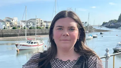 Mared Llywelyn is facing the camera. She has dark hair and is wearing a black dress. Behind her is the Menai Strait. It is a sunny day and there are boats moored in the background.