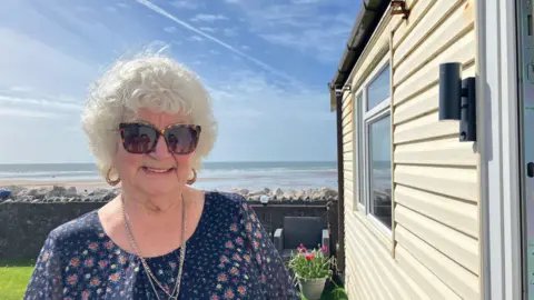 Jennie Dennett/BBC Close-up of Pat Lumb looking directly into the camera. She has grey hair and is wearing sunglasses and a blue top with a floral pattern. She is standing next to her home, which is on the coastline at Walney. The sea and shore can be seen in the background.