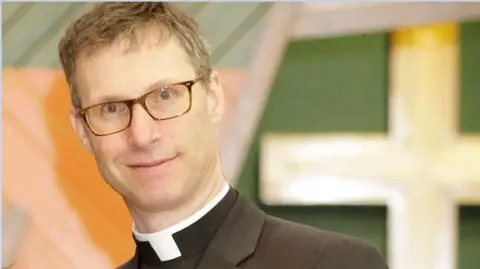 Official diocese portrait of The Right Reverend Philip North in front of a large cross. He is wearing glasses and a dark clerical suit with white collar.