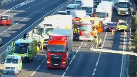 Firefighters and paramedics standing on the four-lane road next to a parked ambulance, fire engine and two lorries. Cones have been used to close the road. Behind them, lorries, vans and cars are queuing.