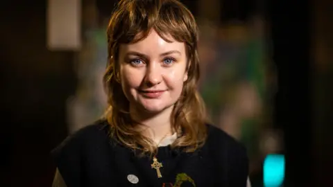 A woman wearing a dark knitted sweater with a cross necklace, standing indoors with soft, colourful, out‑of‑focus lights in the background.