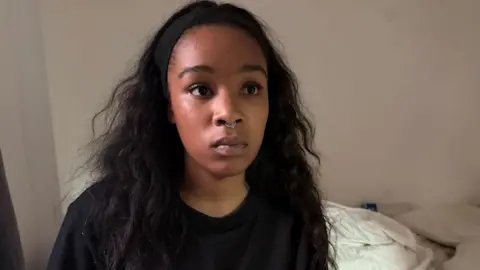 Isabel Rose is seen wearing a black top and a small nose ring, sits facing the camera indoors.