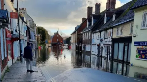DWFRS A man stands on the pavement looking out across the flooded street that stretches in to the distance. Terraced shops line the street and the water comes up to the front doors.