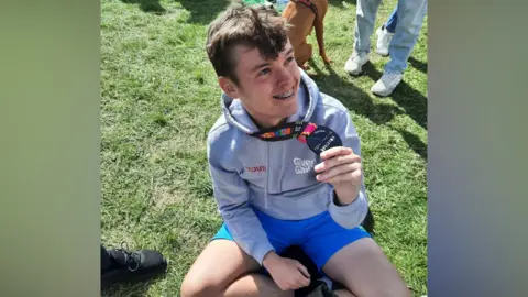 Darren Nunn A young man, Billy Nunn, sits on the ground and holds up his finisher's medal for the Brighton Marathon. He is wearing a light grey top and blue shorts. He has brown hair and braces on his teeth.