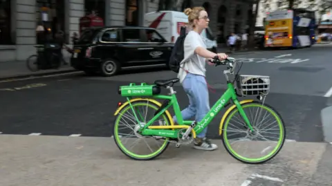A woman wheels a green e-bike across a road in London