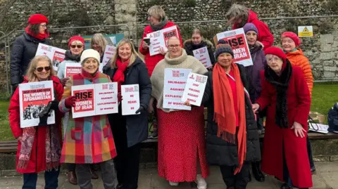 Cad Taylor/BBC A group of women are standing in front of the Abbey walls in Bury St Edmunds holding placards showing support for women in Iran and Afghanistan