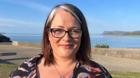 A woman with red hair, with a white streak, and glasses is speaking into a microphone. The background is the sea. She is wearing a floral top.