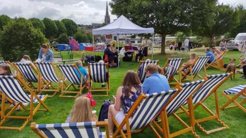 Mole Valley District Council People sit on deckchairs on grass in a public park while a small outdoor band performs under a white canopy, with trees and buildings in the background.