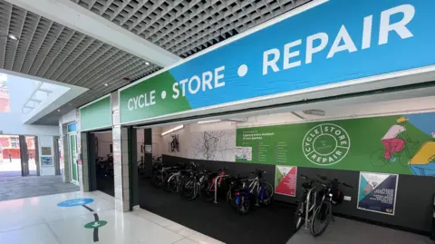 The inside of a shopping centre. A row of bikes are chained up to bike racks in a unit. A blue and green sign above the entrance reads "CYCLE - STORE - REPAIR".