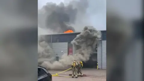 Two firefighters standing in front of an industry unit which has dark smoke coming from the front. There is also an orange blaze coming from the roof.