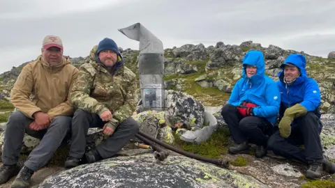 Chris Bullock Espen Thronsen, Chris Bullock, Marianne Rokset and Trond Hjelmtveit are all sitting on rocky ground wearing outdoor weatherproof clothing and hats. Between them is a memorial made from World War Two aeroplane propellers. Attached to it in the middle is a plaque, and beneath it is a photograph of Wilfred Surplice. 