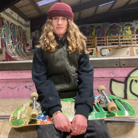 Nottingham skateboarder Miriam Nelson is shown looking straight towards the camera and is holding a skateboard in her lap. 