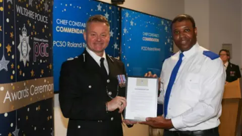 Wiltshire Police Two policemen in a midshot - one in full uniform is giving a certificate to another man wearing a white shirt with blue lapels and blue tie. There are posters in the background saying Wiltshire PCC and 'Chief Constable Commendations. They look like they're on a stage as there's a lectern behind them