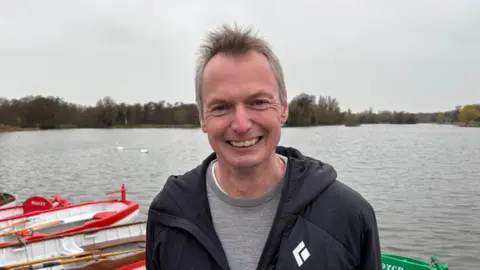 Luke Deal/BBC Hamish Ogilvie smiling at the camera, he is wearing a black coat and gray jumper, behind him are boats on the edge of a lake, with trees and bushes across the water.