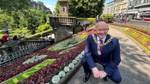 City of Edinburgh Council The Lord Provost is crouching down beside the clock, which is made up of green and purple flowering plants. There are the shops of Princes Street in the background. The Lord Provost is wearing a blue suit, glasses and his badge and chain of office. He is bald and smiling.
