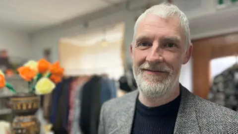 Close-up of Scott McBride with short grey hair and a trimmed beard wearing a textured blazer and dark top, standing indoors in front of a clothing rack and shelves, with a vase of orange and cream flowers visible to the side.