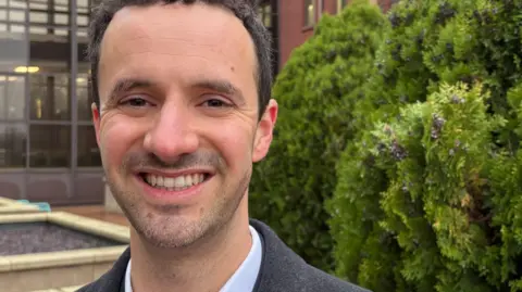 Martin Heath/BBC Chris Hinchliff with short dark hair and slight stubble smiling at the camera and wearing a brown jacket and pink shirt. He is standing in front of small trees to the right, wooden flower beds to the left and the glass and steel facade of a council building behind.
