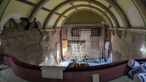 The interior of the Paignton Picture House viewed from the balcony. Two men in high-res jackets and hard hats are at the front looking up. Plaster has come off the barrel roof and there is scaffolding along the front wall. In the front of the balcony are rolls of insulation material.