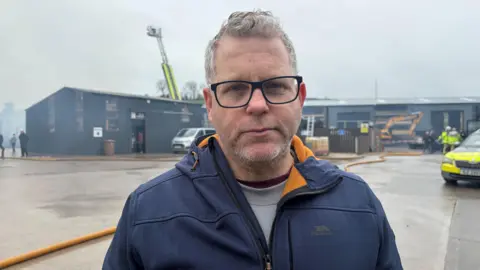 A man if looking into the camera and not smiling. The sky is full of smoke. An aerial ladder is up. A police car, and people in high viz jackets are in the background. 