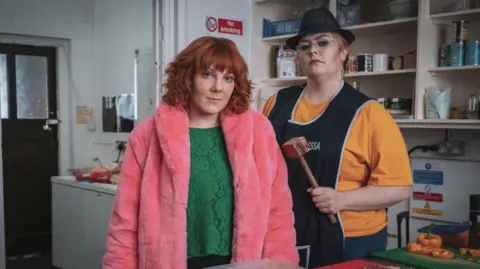 Sophie Willan on the left wearing a green jumper and pink furry jacket, and Jayde Adams on the right wearing a yellow top with an overall. They are standing in front of a shelving unit in a kitchen