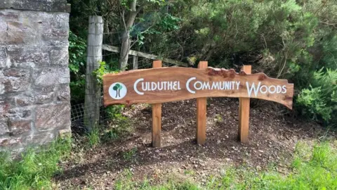 A wooden sign near an entrance to the woods. In white lettering the sign reads: "Culduthel Community Woods". There is a brick wall next to it and vegetation behind it.