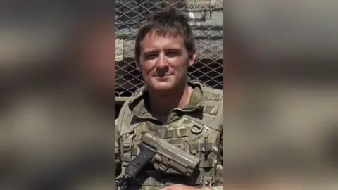 Efrem Brynin A young man in army uniform smiles with a pistol strapped across his chest