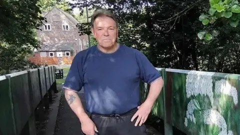 Councillor Phil Millward stands at one of the bridges over Hurley Brook. he is wearing a blue T-shirt and it is sunny. There are trees and a house in the background.