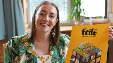 Derry Féile A woman weraing a mayoral chain and a green dress is sat and is smiling. She has long brown hair and is holding a book, with the word feile on its front