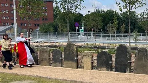 Amanda White/BBC The Right Reverend Dr Eleanor Sanderson walking along, holding a crozier in front of her. She is walking along a narrow path next to a row of gravestones. There are people following her.