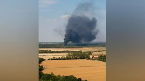 Fountains Media A large plume of dark black smoke emanates from a field fire. The land around it is parched dry.