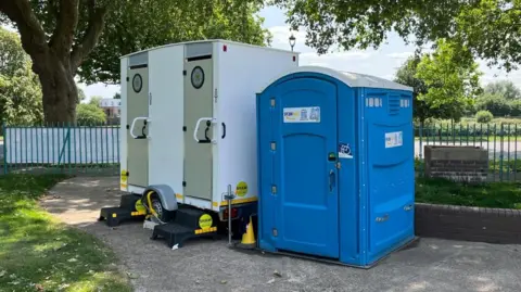 The toilet provision at the Victoria Embankment Paddling Pool, with one blue plastic cabin, and a male and female metal cabin on wheels.