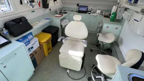 Dentaid A white dentist chair is situated in the middle of the unit surrounded by cupboards and dental equipment. 