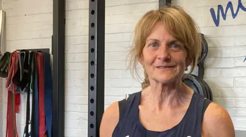 Justin Dealey/BBC A woman stands in a gym in front of a weight rack. She is smiling at the camera. She has blonde hair part of which has been tied back. She wears a navy blue vest with a Team GB logo on it as well as an Adidas logo.