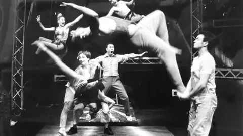 A group of physical theatre performers jumping, doing acrobatics and performing aerial manoeuvres. Rigging can be seen around them. It is an old black and white photograph.