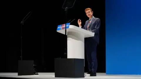 PA Media Andy Street in a blue suit stands gesticulating at a podium