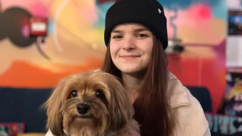 A Hayanese dog with golden fur and large eyes is being held by her owner who is wearing a black woolly hat and has long brown hair. The woman has a nose ring and is smiling at the camera.