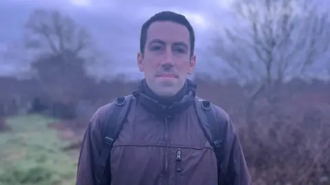 A man with dark hair and a brown jacket stands with a black backpack on. In the background is a green area with trees.