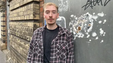 Huw has short blonde hair and brown stubble with a brown moustache. He is wearing a purple, white and grey plaid shirt with a grey top underneath. He is standing in front of a portion of wall that has graffiti on it.