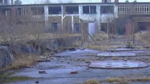Robert Wade/Geograph The deserted site of Huncoat power station. It is a long two-storey brick building with a taller section to the left which have been vandalised. To the front are overgrown flat areas with covers on the ground and sprouting weeds 