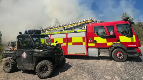 A small black vehicle with the fire service logo on the side. On the back of its trailer it has yellow pipes. Next to it is a red and yellow fire engine with a metal grey ladder on top. There are shadows of firemen inside the vehicle.