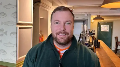 Jack Hannam has short brown hair and a goatee beard and moustache. He is smiling and wearing a green fleece-like top and an orange and blue top underneath. He is standing behind the bar and there are beer taps behind him.