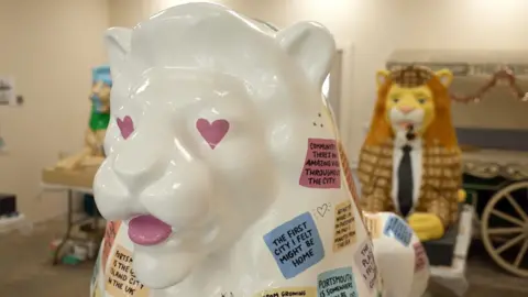 The image shows three lion sculptures. Two in the background are slightly blurred and the one if the foreground is in focus. The one in the front is white and painted with pink heart eyes. It has a number of different coloured squares which each say different messages about Portsmouth. The one in the background is painted in to look like it is wearing a suit. It has a brown jacket on and a black tie. It has a ginger mane and a brown hat on. The third lion is inspired by history of libraries. It is a brown lion, which has green and grey on it.