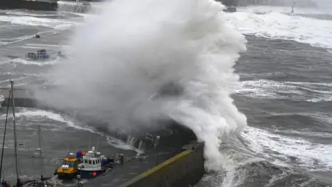 PA Media A massive wave blows over the harbour wall at Stonehaven. The plume of water is white, above a grey sea. Two small boats are in the harbour in the foreground.