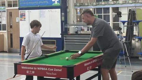 Andrea Pluck/BBC A child in a white polo top holding a cue receives instruction from Kyren Wilson as they stand over a small snooker table.