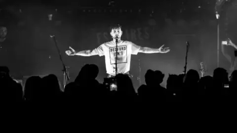 Neil McCarty Tom Grennan performing at Esquires in Bedford. The picture is black and white and Grennan is in front of a microphone with his arms stretched out.