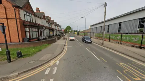 A generic image of Aston Lane, going through the middle of the photo, from the bottom going upwards. Two cars are on the road. Housing is to the left of the street and another building is to the right.
