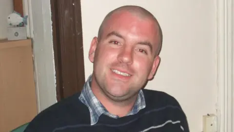 Norfolk Police A smiling man wearing a shirt with a dark striped jumper on top